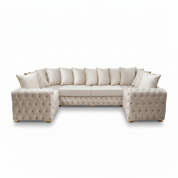 A large tufted sectional sofa in off-white/cream upholstery arranged in an L-shape in a styled living room. The sofa is adorned with multiple matching throw pillows and sits on a plush light beige rug, with wall sconces, a framed abstract painting, curtains, and a potted palm in the background.